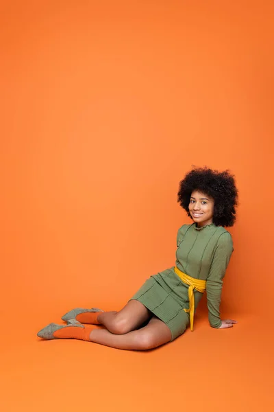Full length of positive african american teenager with bold makeup and green dress smiling at camera while sitting on orange background, youth culture and generation z concept — Stock Photo