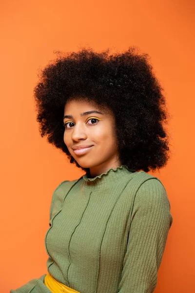 Retrato de la muchacha afroamericana adolescente de moda con maquillaje audaz usando vestido verde y sonriendo a la cámara aislada en naranja, cultura juvenil y el concepto de generación z - foto de stock