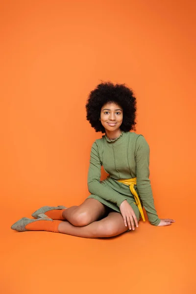 Modèle afro-américain sur toute la longueur avec un maquillage audacieux portant une robe verte tout en étant assis et en regardant la caméra sur fond orange, la culture des jeunes et le concept de génération z — Photo de stock