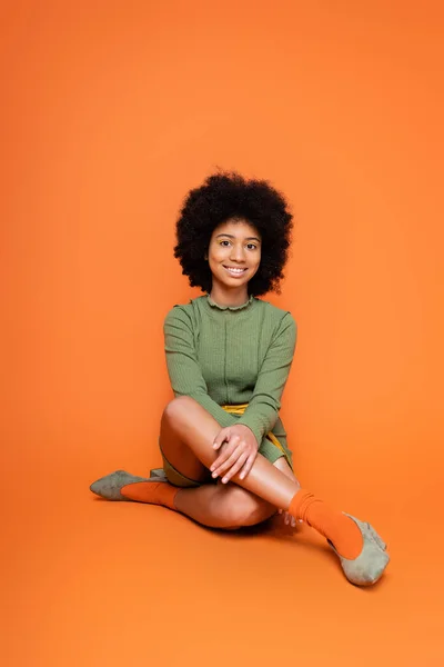 Comprimento total de alegre modelo americano africano adolescente com maquiagem colorida vestindo vestido verde e sentado em fundo laranja, cultura juvenil e conceito z geração — Fotografia de Stock
