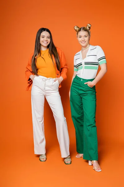 Full length of cheerful and confident brunette teenager holding hands in pockets while posing near blonde girlfriend on orange background, fashionable girls with sense of style, friendship — Stock Photo
