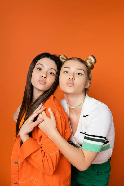 Retrato de namoradas adolescentes elegantes com maquiagem colorida soprando beijo de ar na câmera e abraçando enquanto estão juntas isoladas em laranja, meninas da moda com senso de estilo — Fotografia de Stock