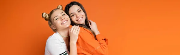 Positive and trendy blonde teenage model with bright makeup hugging brunette girlfriend and looking at camera isolated on orange, fashionable girls with sense of style, banner with copy space — Stock Photo