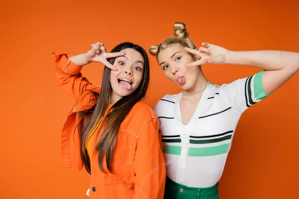 Portrait of trendy teenage girlfriends with bright makeup sticking out tongues and showing peace gesture at camera while standing isolated on orange, fashionable girls with sense of style — Stock Photo
