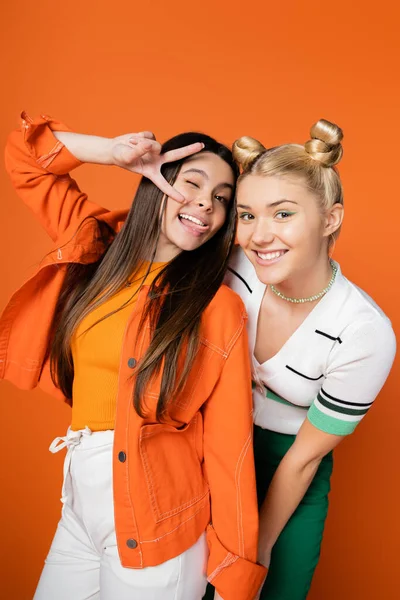 Cheerful blonde and brunette teenage girlfriends with bright makeup and stylish outfits gesturing and posing at camera while standing isolated on orange, fashionable girls with sense of style — Stock Photo