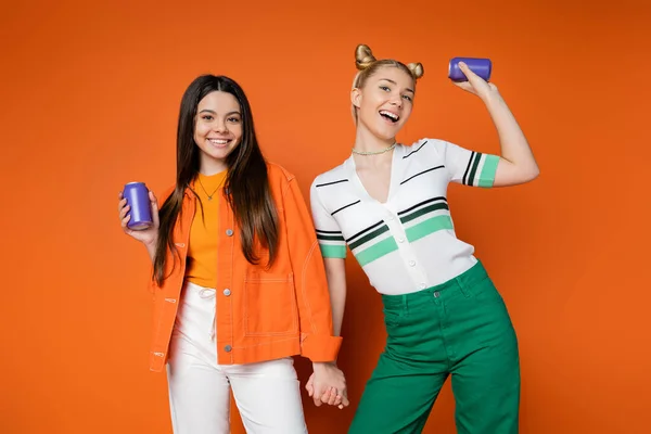 Cheerful blonde and brunette teenage girlfriends in casual outfits holding drink in tin cans and holding hands while standing on orange background, fashionable girls with sense of style — Stock Photo