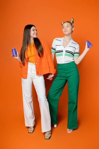 Full length of smiling and stylish brunette teenager holding hand of blonde girlfriend and drink in tin can and standing together on orange background, fashionable girls with sense of style — Stock Photo