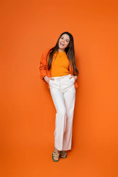 Full length of cheerful and brunette teenage girl in stylish casual outfit holding hands in pockets and looking at camera while standing on orange background, cool and confident teenage girl — Stock Photo