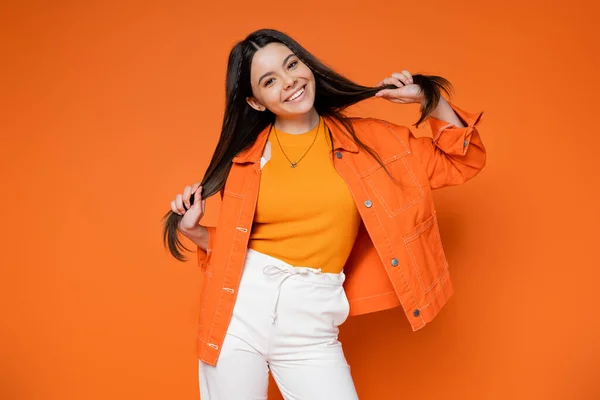 Trendy brunette teenage model in denim jacket and pants touching hair and smiling at camera while standing on orange background, cool and confident teenage girl — Stock Photo