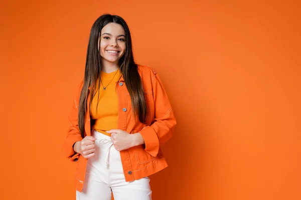 Fashionable and brunette teenage girl in denim jacket and pants smiling while looking at camera and posing on orange background, cool and confident teenage girl — Stock Photo