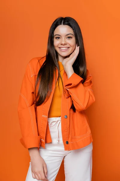 Porträt eines fröhlichen und brünetten Teenie-Modells in Jeansjacke und Hose, das Gesicht berührt und in die Kamera schaut, während es isoliert auf einem orangefarbenen, coolen und selbstbewussten Teenager-Mädchen steht — Stockfoto
