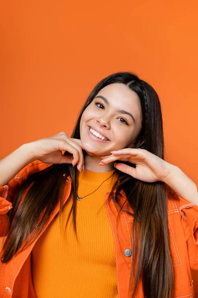 Portrait of positive and stylish brunette teenage model in denim jacket touching chin and smiling at camera while standing isolated on orange, cool and confident teenage girl — Stock Photo