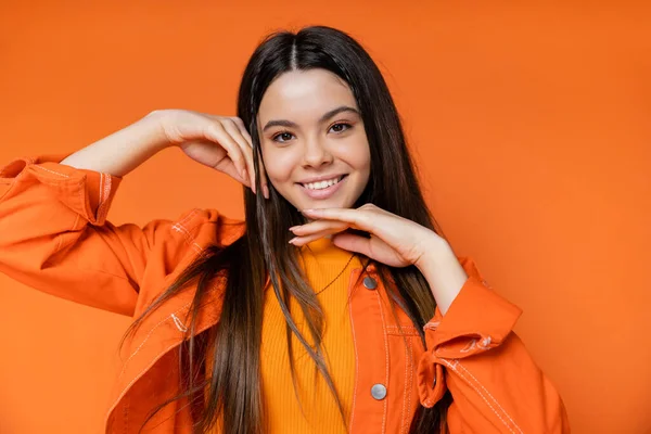 Porträt eines trendigen und brünetten Teenie-Modells in Jeansjacke, das Gesicht berührt und in die Kamera schaut, während es isoliert auf einem orangefarbenen, coolen und selbstbewussten Teenager-Mädchen posiert — Stockfoto