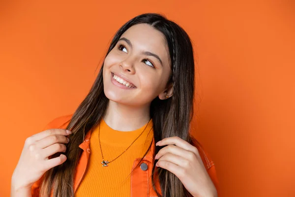 Porträt eines fröhlichen Teenie-Models in Jeansjacke, das brünettes Haar berührt und wegschaut, während es isoliert auf einem orangefarbenen, coolen und selbstbewussten Teenager-Mädchen posiert — Stockfoto