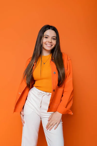 Cheerful and brunette teenage girl with bright makeup wearing trendy outfit while standing and posing on orange background, cool and confident teenage girl — Stock Photo