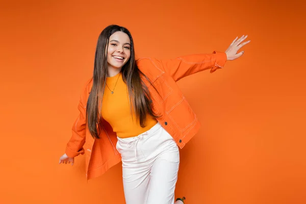 Fashionable and cheerful brunette teenage girl in denim jacket and pants looking at camera while posing isolated on orange, cool and confident teenage girl, gen z fashion — Stock Photo