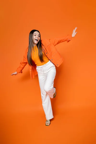 Full length of excited and trendy brunette teen model in denim jacket and pants posing and looking at camera while standing on orange background, cool and confident teenage girl — Stock Photo