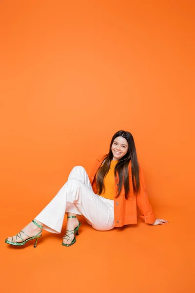 Full length of cheerful and fashionable brunette teenager with bold makeup wearing heels and denim jacket while looking at camera and sitting on orange background, cool and confident teenage girl — Stock Photo
