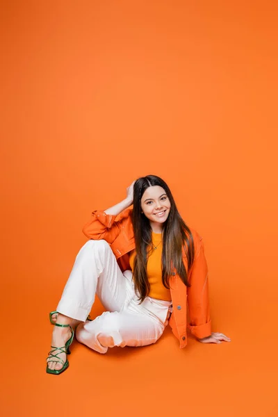 Chica adolescente morena de moda en chaqueta de mezclilla, pantalones y tacones posando y sonriendo a la cámara mientras está sentado en fondo naranja, chica adolescente fresca y segura de sí misma, gen z moda - foto de stock