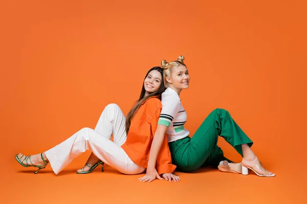 Full length of fashionable teenage girlfriends in casual outfits and heels smiling at camera while sitting back to back on orange background, cool and confident teenage girls — Stock Photo