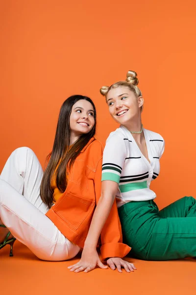 Joyful and trendy blonde and brunette teenage girlfriends with bold makeup smiling and sitting back to back and looking at each other on orange background, cool and confident teenage girls — Stock Photo
