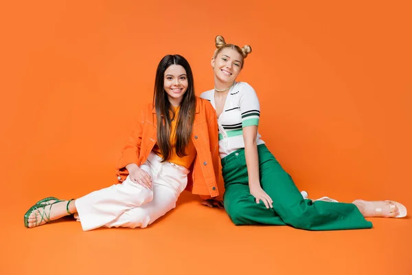 Volle Länge der modischen Teenager-Freundinnen mit hellem Make-up tragen lässige Outfits, während sie nebeneinander sitzen und in die Kamera auf orangefarbenem Hintergrund schauen, coole und selbstbewusste Teenager-Mädchen — Stockfoto