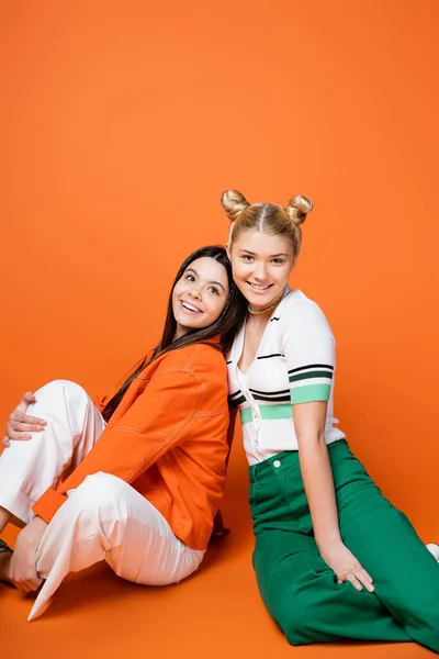 Positive and stylish blonde teenage model in casual outfit smiling at camera while sitting next to brunette girlfriend on orange background, cool and confident teenage girls — Stock Photo