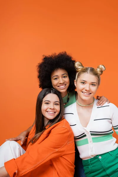 Lächelndes afrikanisch-amerikanisches Teenager-Mädchen mit hellem Make-up umarmt stilvolle Freundinnen in lässigen Outfits und posiert gemeinsam isoliert auf orangefarbenen, coolen und selbstbewussten Teenager-Mädchen — Stockfoto