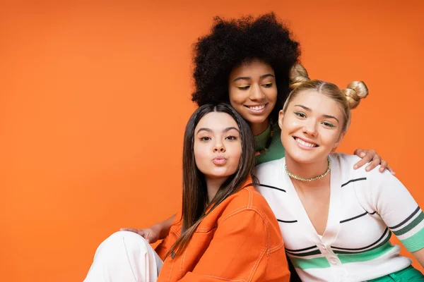 Fröhliche afrikanisch-amerikanische Teenager-Mädchen mit hellem Make-up umarmt stilvolle blonde und brünette Freundinnen, während sie zusammen isoliert auf orangefarbenen, coolen und selbstbewussten Teenager-Mädchen posieren — Stockfoto