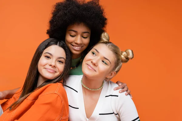 Lächelndes afrikanisch-amerikanisches Teenie-Model mit hellem Make-up, das modische Freundinnen umarmt und isoliert auf orangefarbenen, coolen und selbstbewussten Teenagermädchen posiert, diverse Rassen — Stockfoto