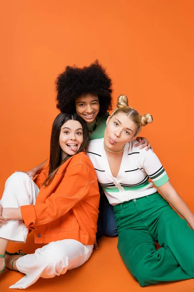 Positive und multiethnische Teenager-Freundinnen in stilvollen lässigen Outfits, die die Zunge herausstrecken und in die Kamera zwinkern, während sie auf orangefarbenem Hintergrund posieren, coole und selbstbewusste Teenager-Mädchen, diverse Rassen — Stockfoto