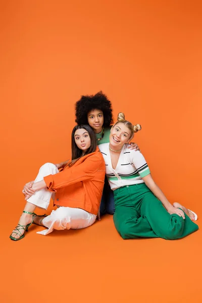 Comprimento total de namoradas adolescentes alegres e multiétnicas em roupas casuais na moda posando juntas e olhando para a câmera no fundo laranja, meninas adolescentes multiculturais legais e confiantes — Fotografia de Stock