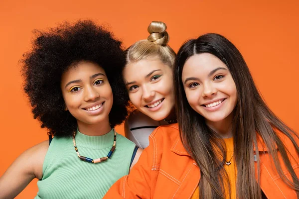 Portrait of fashionable and multiethnic teenage models with colorful makeup smiling at camera and standing isolated on orange, cool and confident multicultural teenage girls — Stock Photo