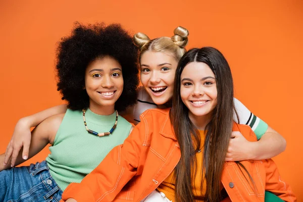 Joyful blonde teenage girl hugging fashionable and multiethnic girlfriends with bold makeup while standing isolated on orange, cool and confident multicultural teenage girls, diverse races — Stock Photo