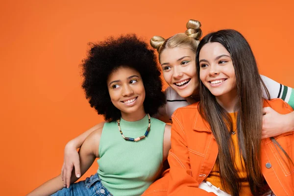Adolescente loira alegre com maquiagem ousada abraçando namoradas inter-raciais na moda e olhando juntos isolado em laranja, cool e confiante multicultural adolescentes meninas — Fotografia de Stock