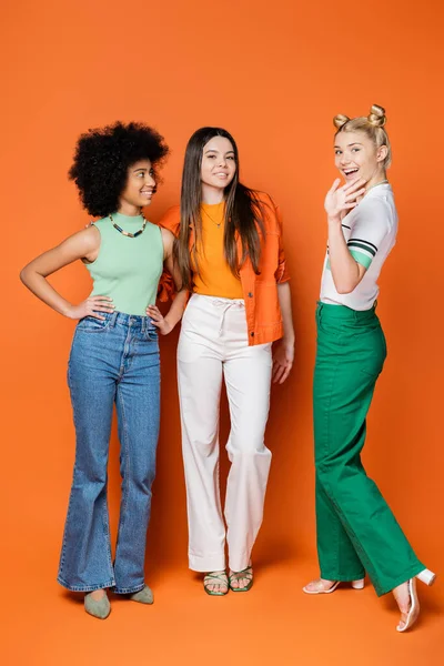 Full length of stylish blonde teenager waving hand at camera while standing near trendy and multiethnic girlfriends on orange background, cool and confident multicultural teenage girls — Stock Photo