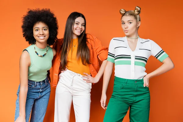 Adolescentes alegres e multiétnicos com maquiagem ousada posando em roupas da moda e olhando para a câmera juntos no fundo laranja, meninas adolescentes multiculturais legais e confiantes — Fotografia de Stock