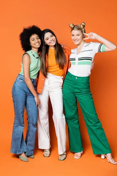 In voller Länge positiver blonder Teenager, der Siegesgeste zeigt, während er mit stilvollen multiethnischen Freundinnen posiert und auf orangefarbenem Hintergrund steht, coole und selbstbewusste multikulturelle Teenager-Mädchen — Stockfoto