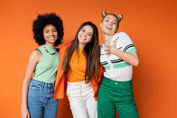 Smiling blonde teenager in casual outfit hugging interracial girlfriends with bold makeup and looking at camera on orange background, trendy outfits and fashion-forward looks — Stock Photo