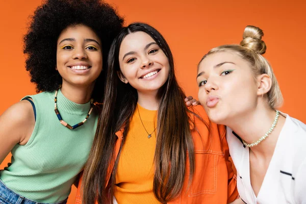 Cheerful and multiethnic teen girlfriends with bold makeup and in trendy casual clothes looking at camera while blonde friend pouting lips isolated on orange, trendy outfits and fashion-forward looks — Stock Photo