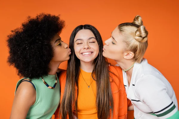 Teen african american and blonde girlfriends with colorful makeup kissing cheerful and stylish girlfriend while standing isolated on orange, trendy outfits and fashion-forward looks — Stock Photo