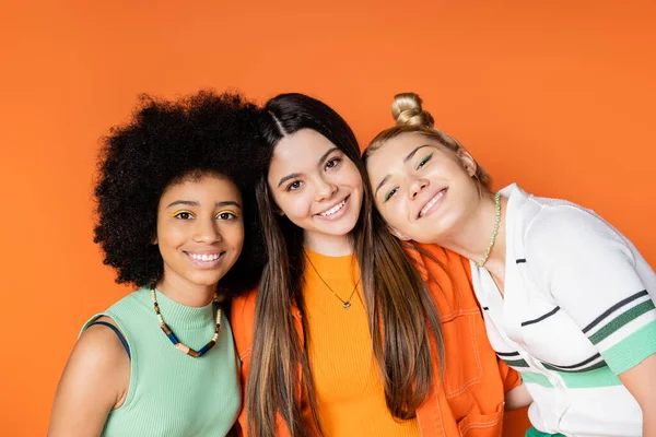 Portrait of smiling and multiethnic teen girlfriends with bright makeup looking at camera while posing together isolated on orange, trendy outfits and fashion-forward looks, diverse races — Stock Photo