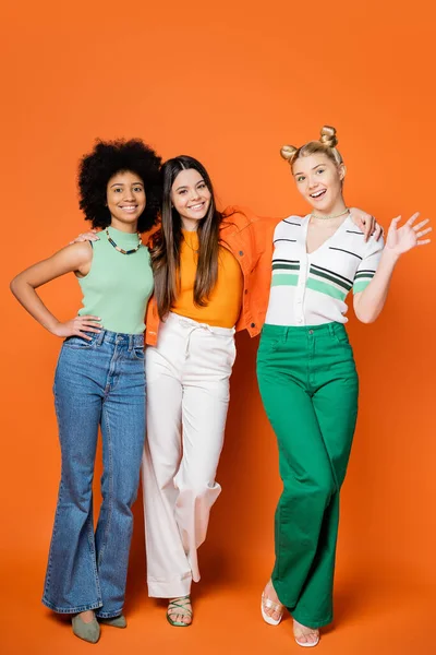 Lächelndes und stilvolles blondes Teenie-Mädchen, das in die Kamera winkt, während es neben multiethnischen Freundinnen steht, mit hellem Make-up auf orangefarbenem Hintergrund, trendigen Outfits und modisch zukunftsweisenden Blicken — Stockfoto