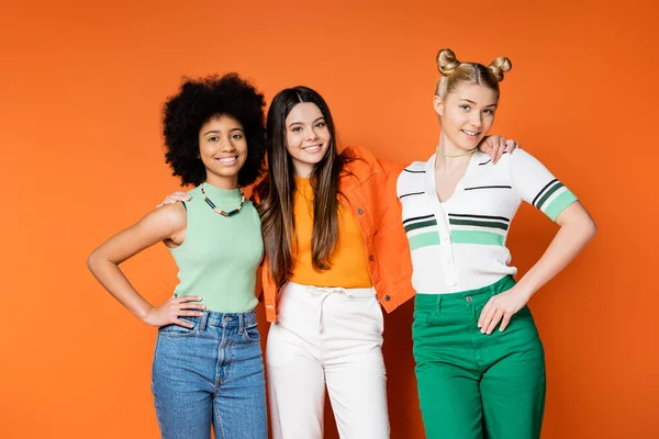 Cheerful brunette teenager in stylish casual outfit hugging interracial friends with bright makeup and standing on orange background, trendy outfits and fashion-forward looks — Stock Photo