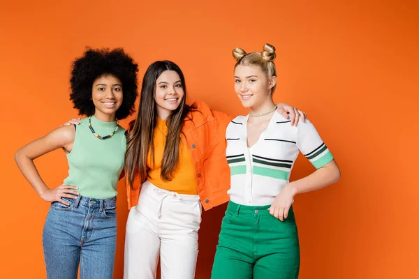 Lächelndes brünettes Mädchen in lässigem Outfit umarmt gemischtrassige Freundinnen mit trendigem, hellem Make-up und posiert auf orangefarbenem Hintergrund, trendigen Outfits und modisch zukunftsweisenden Blicken — Stockfoto