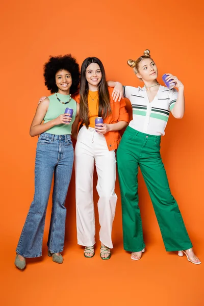 Full length of cheerful and stylish teenager hugging multiethnic girlfriends and holding canned drink while posing together on orange background, trendy outfits and fashion-forward looks — Stock Photo
