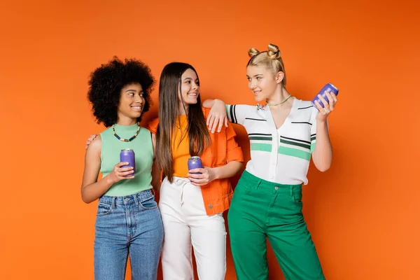 Позитивный блондинка подросток в стильном наряде держа напиток в жестяной банке и глядя на многоэтнических девушек в повседневных нарядах на оранжевом фоне, модные наряды и моды вперед взгляды — стоковое фото