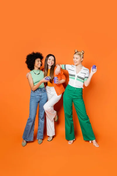 Full length of positive and multiethnic teenage girlfriends in stylish outfits holding canner drink and posing on orange background, trendy outfits and fashion-forward looks — Stock Photo