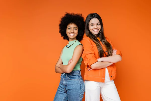 Stylish and fashionable multiethnic teen girlfriends in casual outfits crossing arms and standing back to back isolated on orange, teen fashionistas with impeccable style concept, diverse races — Stock Photo