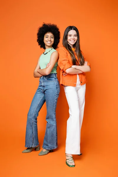 Full length of smiling and multiethnic teenage girlfriends in trendy casual outfits crossing arms and standing on orange background, teen fashionistas with impeccable style concept — Stock Photo